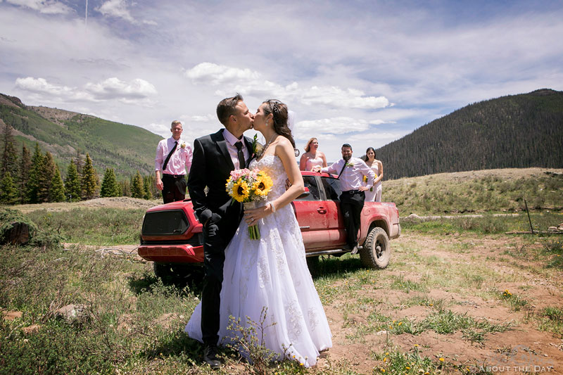 Bride and Groom kiss in front of 4x4 with wedding party in it