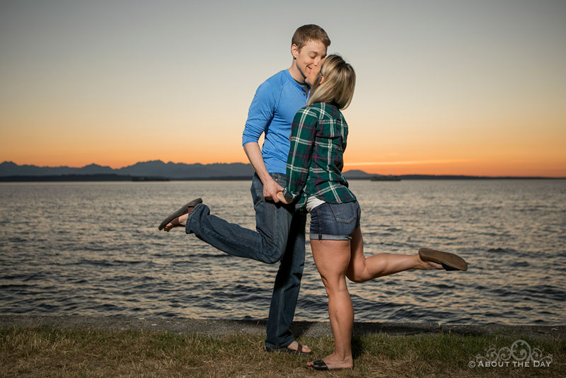 Andrew and Alex lift a foot while kissing at sunset at Alki Beach